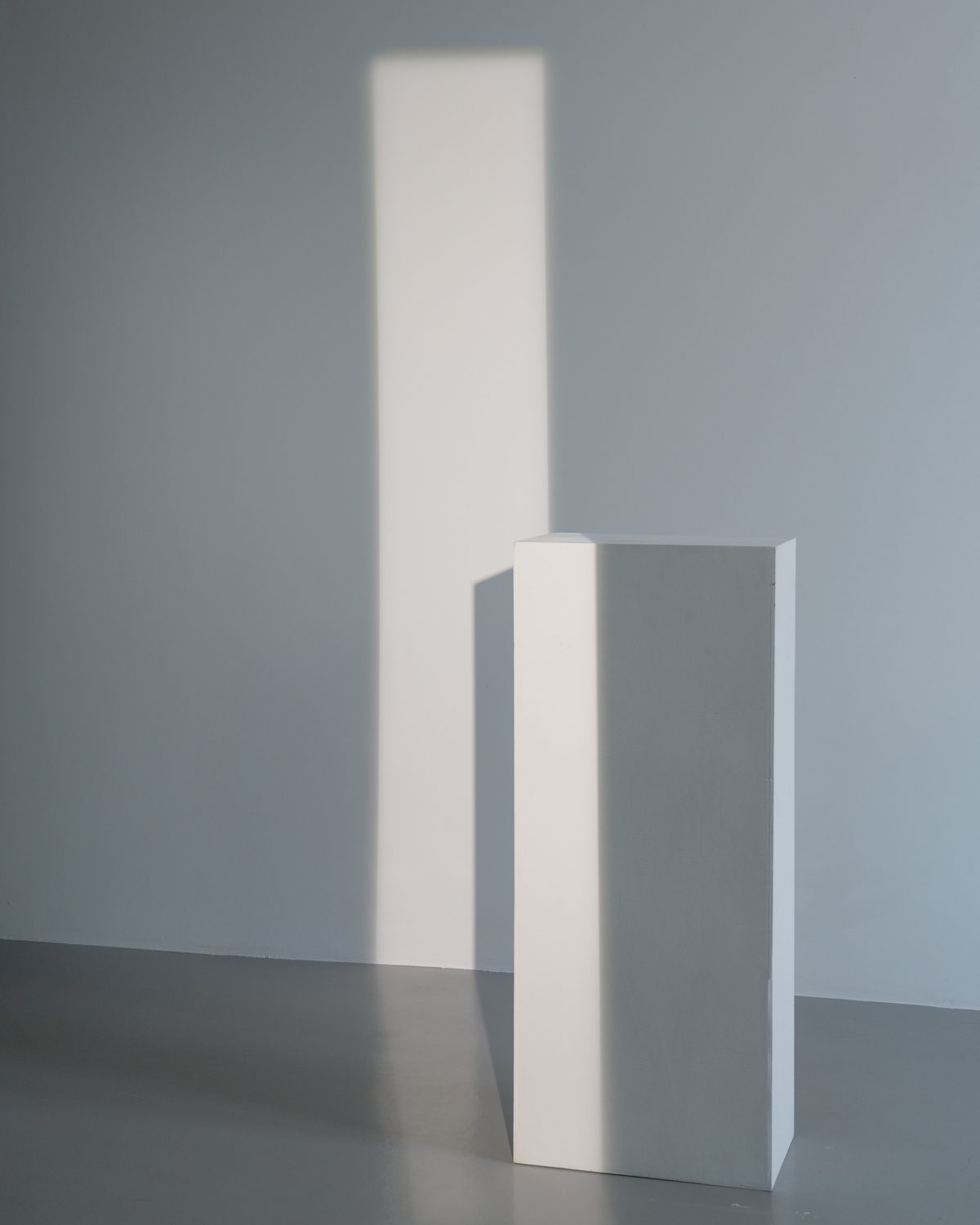 Een lege witte presentatiezuil in een lichte ruimte, met een streep zonlicht op de muur erachter